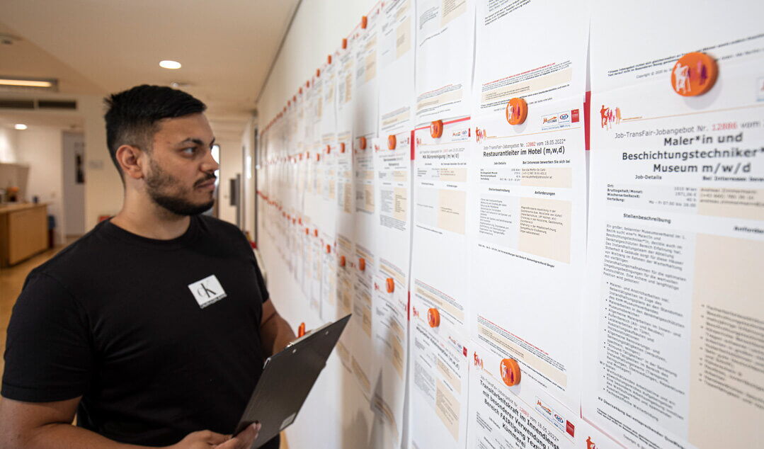 Ein junger Mann mit Dreitagebart und schwarzem T-Shirt schaut konzentriert auf ein Jobangebot , das zusammen mit zahlreichen weiteren Stellenanzeigen an einer Wand im Job-TransFair-Büro angebracht ist. Er hält ein Klemmbrett in der Hand.