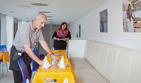 Ein Herr mit Kellner-Schürze deckt unter Anleitung einer Dame mit Notizzettel Tische ein. Er ist Transitarbeitskraft in der Kümmerei und absolviert ein Training-on-the-Job in der Kantine Vier Zehn.
