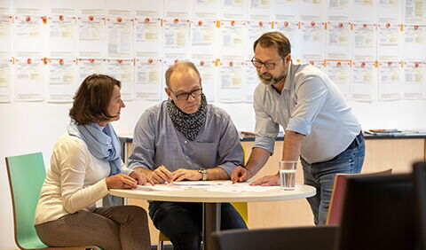 Drei Personen sitzen an einem runden Tisch im Job-TransFair-Büro und tauschen sich über das Stellenangebot aus.