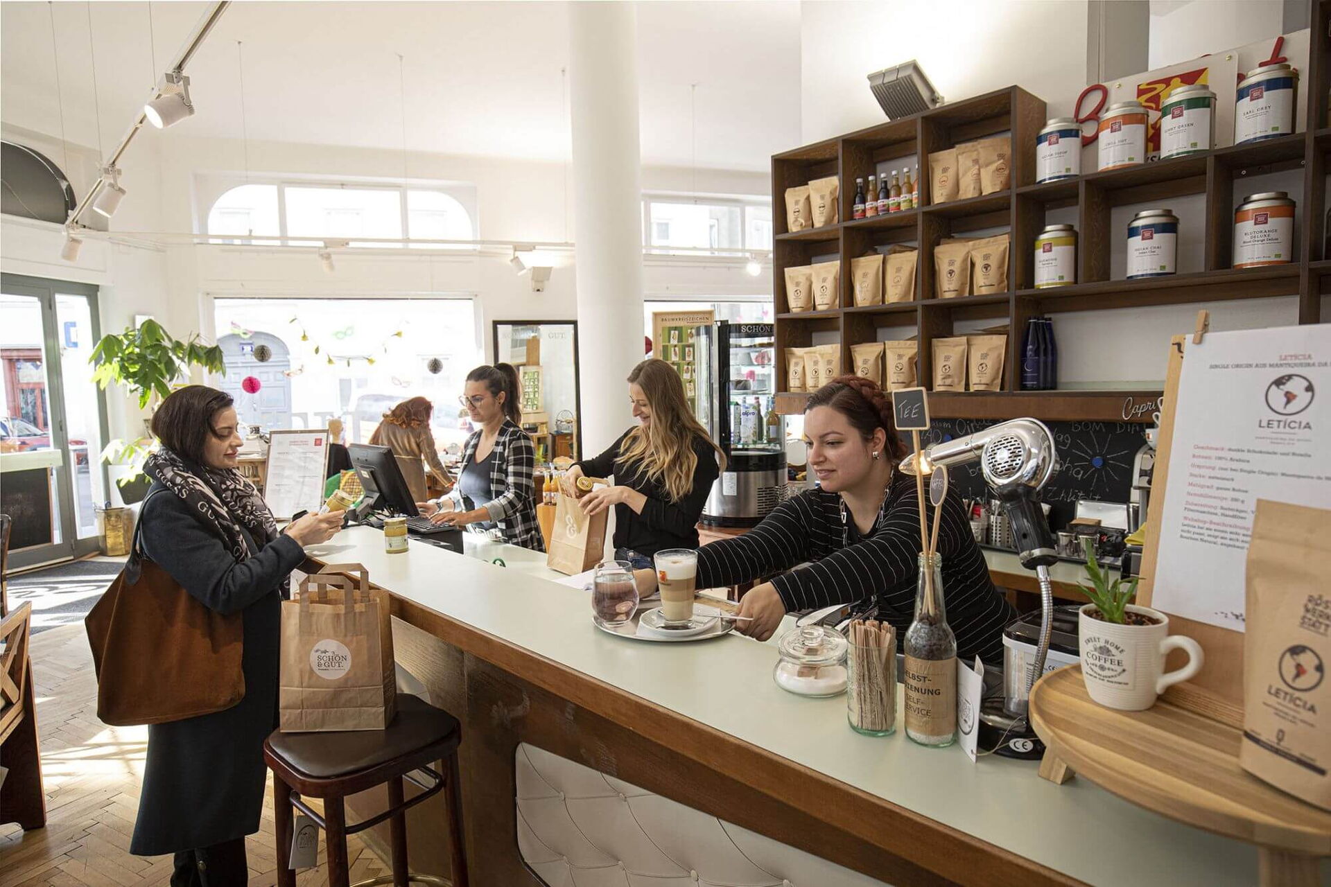 In unserem Social Concept Store Schön und Gut bereiten unsere Mitarbeiterinnen einen Kaffee und gekaufte Waren einer Kundin an der Theke vor.