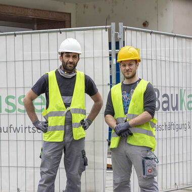 Zwei Personen posieren vor dem "Baukarussell"-Transparent für ein Foto.