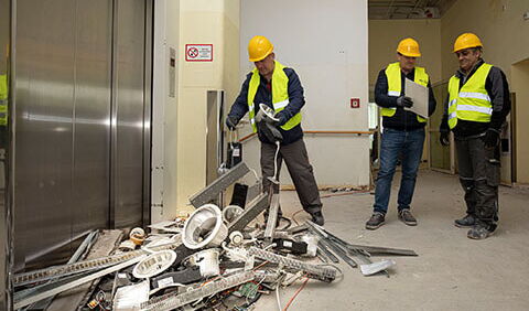 Ein Bauarbeiter des Kümmerei-Teams sortiert wieder verwendbare metallische Bauteile eines Abbruchgebäudes. Sein Teamleiter und ein Kollege stehen hinter ihm.