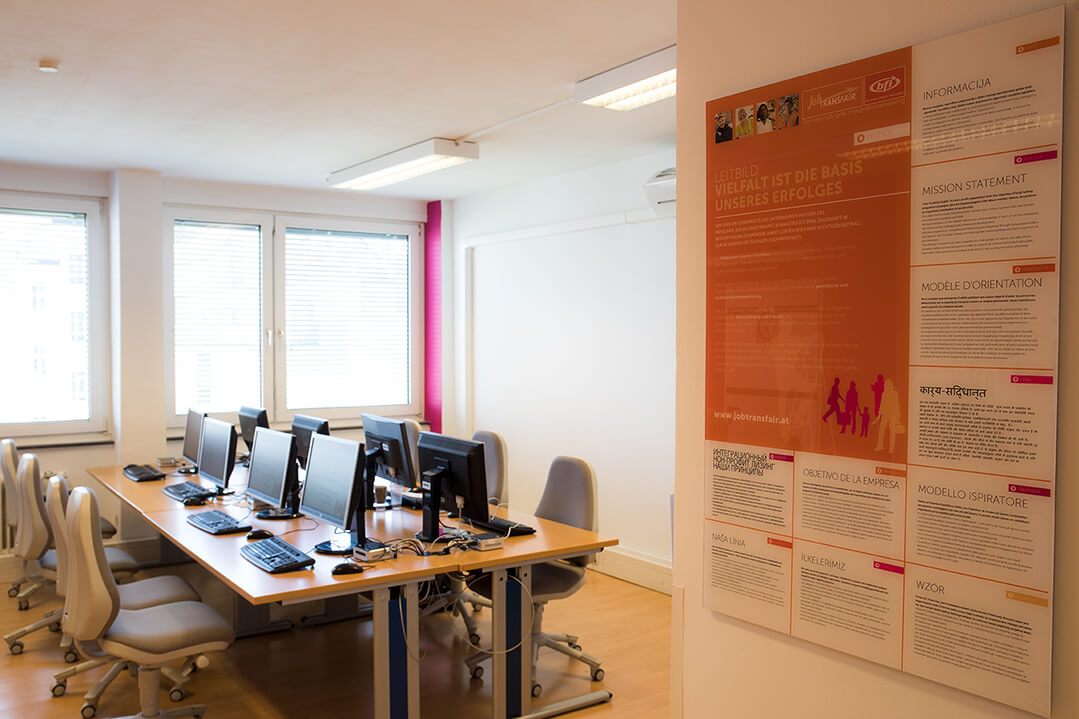 Blick ins Job-TransFair Bewerbungsbüro. Zwei einander zugewandte Reihen mit PC-Arbeitsplätzen. Rechts im Bild sieht man ein Plakat, auf dem das Leitbild in 10 Sprachen übersetzt wurde.