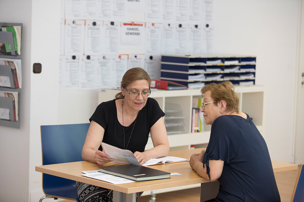Zwei Frauen sitzen sich an einem rechteckigen Tisch gegenüber. Die Beraterin, links im Bild präsentiert der Teilnehmerin, rechts im Bild, den Projektfolder Terra für ältere Migrantinnen und Migranten.