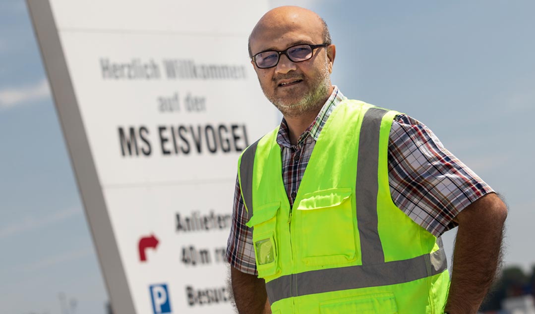 Ein Mann über 50 mit kariertem Kurzarmhemd und gelber Warnweste blickt selbstbewusst in die Kamera. Hinter ihm ragt ein Schild mit dem Text Willkommen auf der MS Eisvogel" auf.