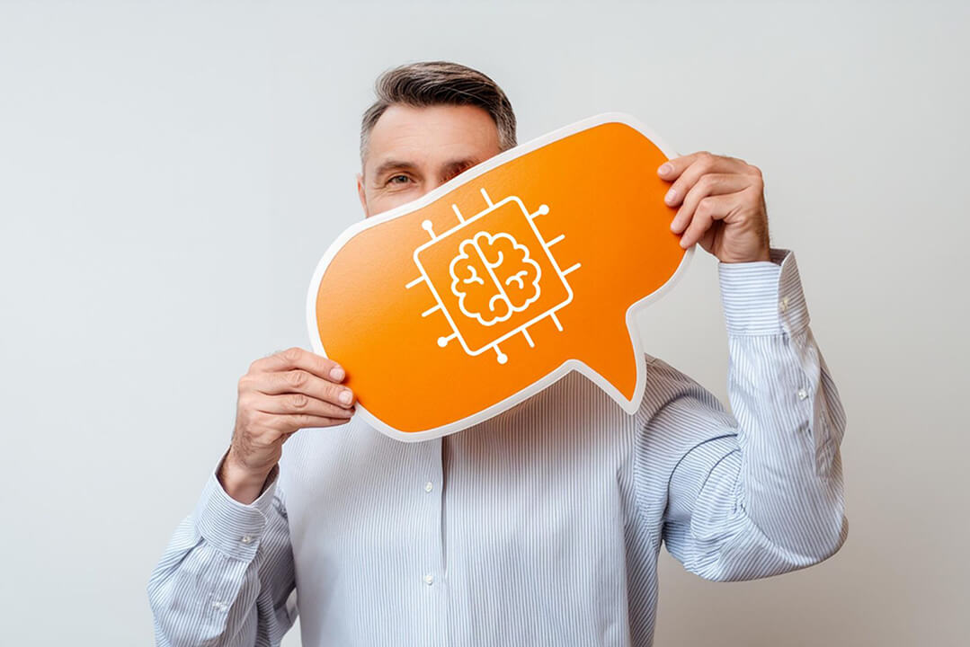 Ein modern gekleideter Mann ist halbnah zu sehen. Er hält ein oranges Schild in der Hand, das die obere Hälfte seines Gesichts verdeckt. Das Schild zeigt das Piktogramm eines Gehirns, das in einem Behälter steckt und Künstliche Intelligenz symbolisiert.