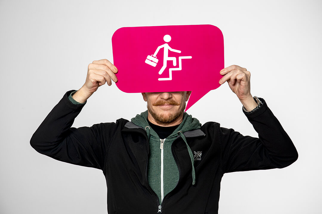 Ein modern gekleideter Mann mit Vollbart ist in Nahaufnahme zu sehen. Er hält ein pinkes Schild in der Hand, das die obere Hälfte seines Gesichts verdeckt. Das Schild zeigt das Piktogramm eines Mannes mit Arbeitstasche, der drei Stufen nach oben nimmt.