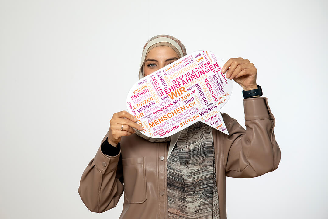 Eine junge Frau mit modischem Kopftuch und beiger Lederjacke hält ein Schild in der Hand, das das Leitbild als bunte Wortwolke abbildet.