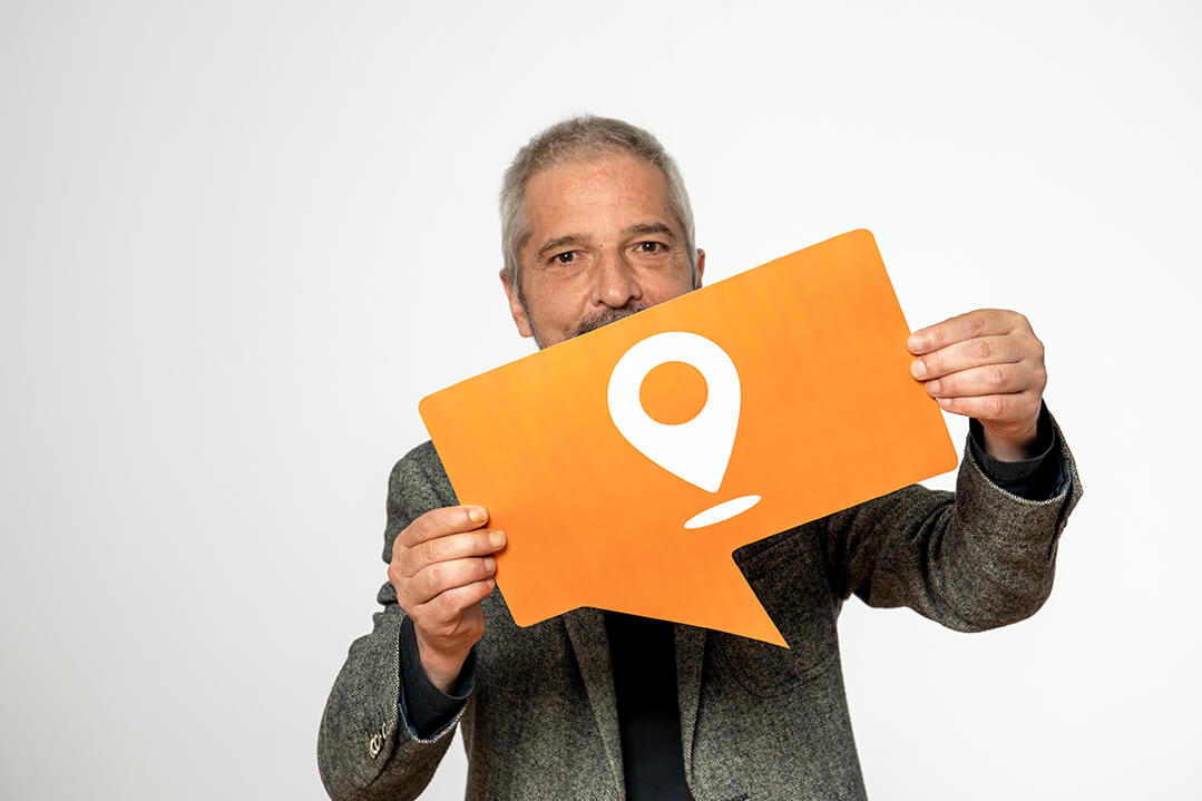 Ein Mann mit grauen kurzen Haaren, Dreitagebart und Sakko hält ein oranges Schild in Form einer Sprechblase in der Hand. Es zeigt ein Piktogramm mit einem Pfeil, der das Wort Hier darstellt.