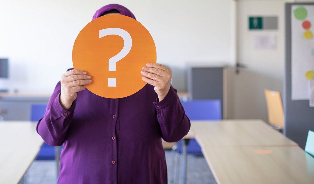 Eine Dame in einem weiten violetten Kleid und Kopftuch hält ein oranges kreisförmiges Schild mit einem Fragezeichen vor ihrem Kopf.