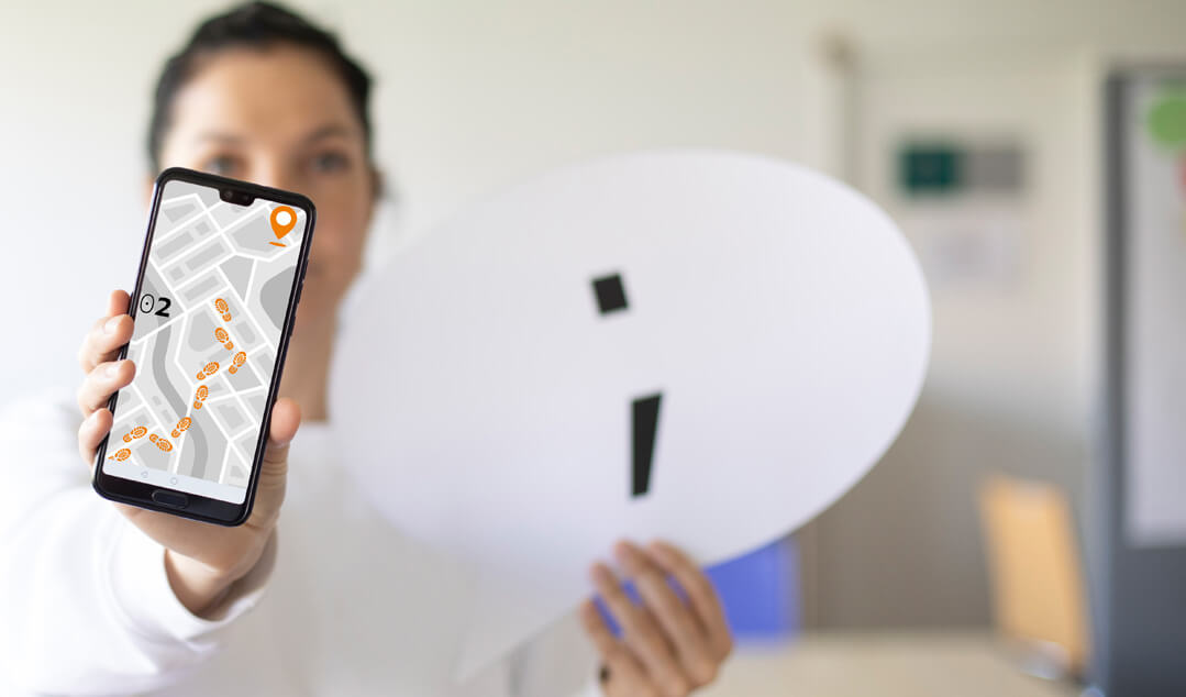 Eine Dame in einem weißen Hemd hält ein Handy mit einem Routenplan sowie ein weißes Schild in Form einer Sprechblase in die Kamera.