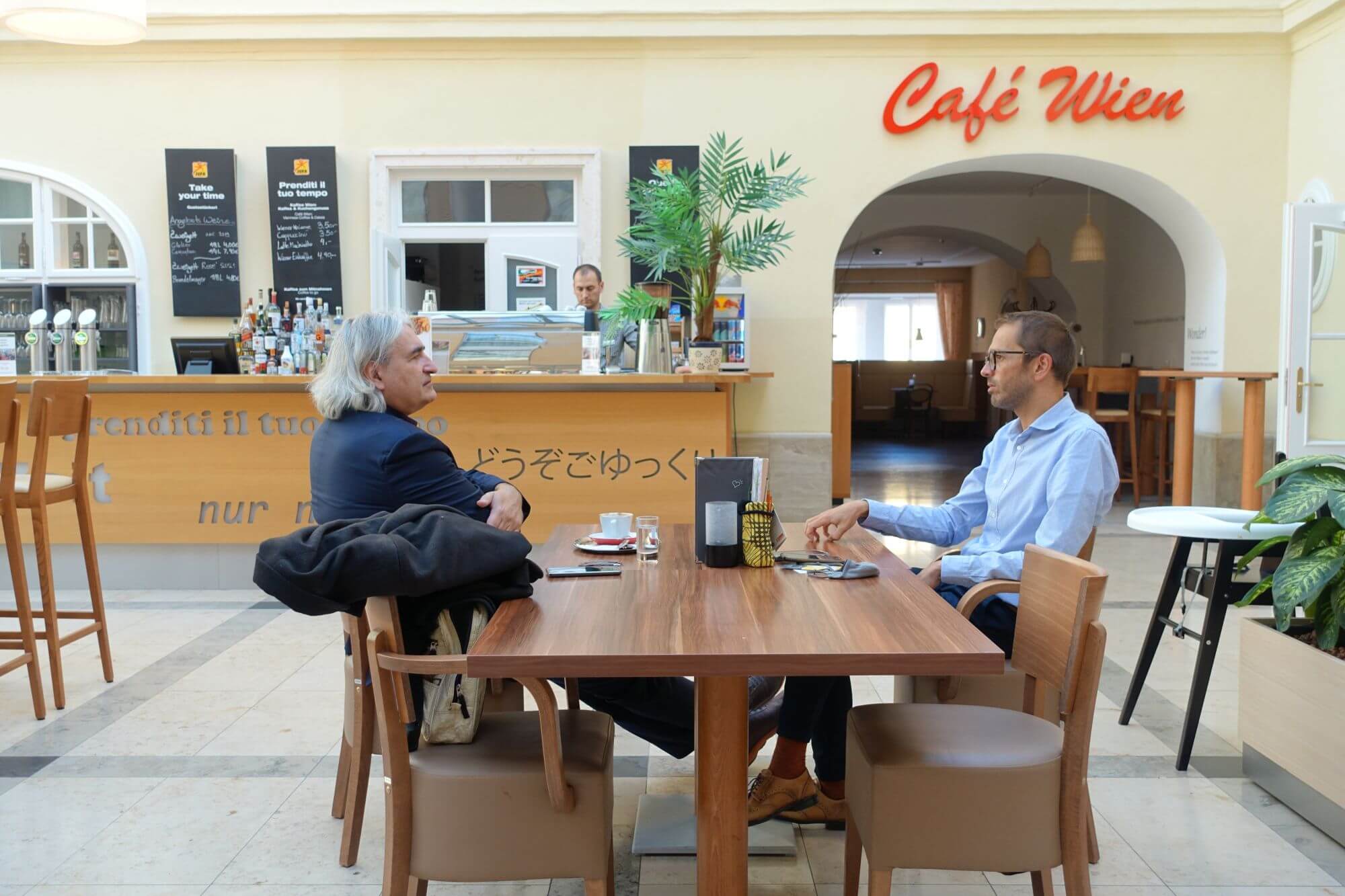 Sven Winter und Thomas Rihl sitzen im Gespräch an einem Tisch im großen Vestibül des Jufa Hotel Wien City. Im Hintergrund die Bar.