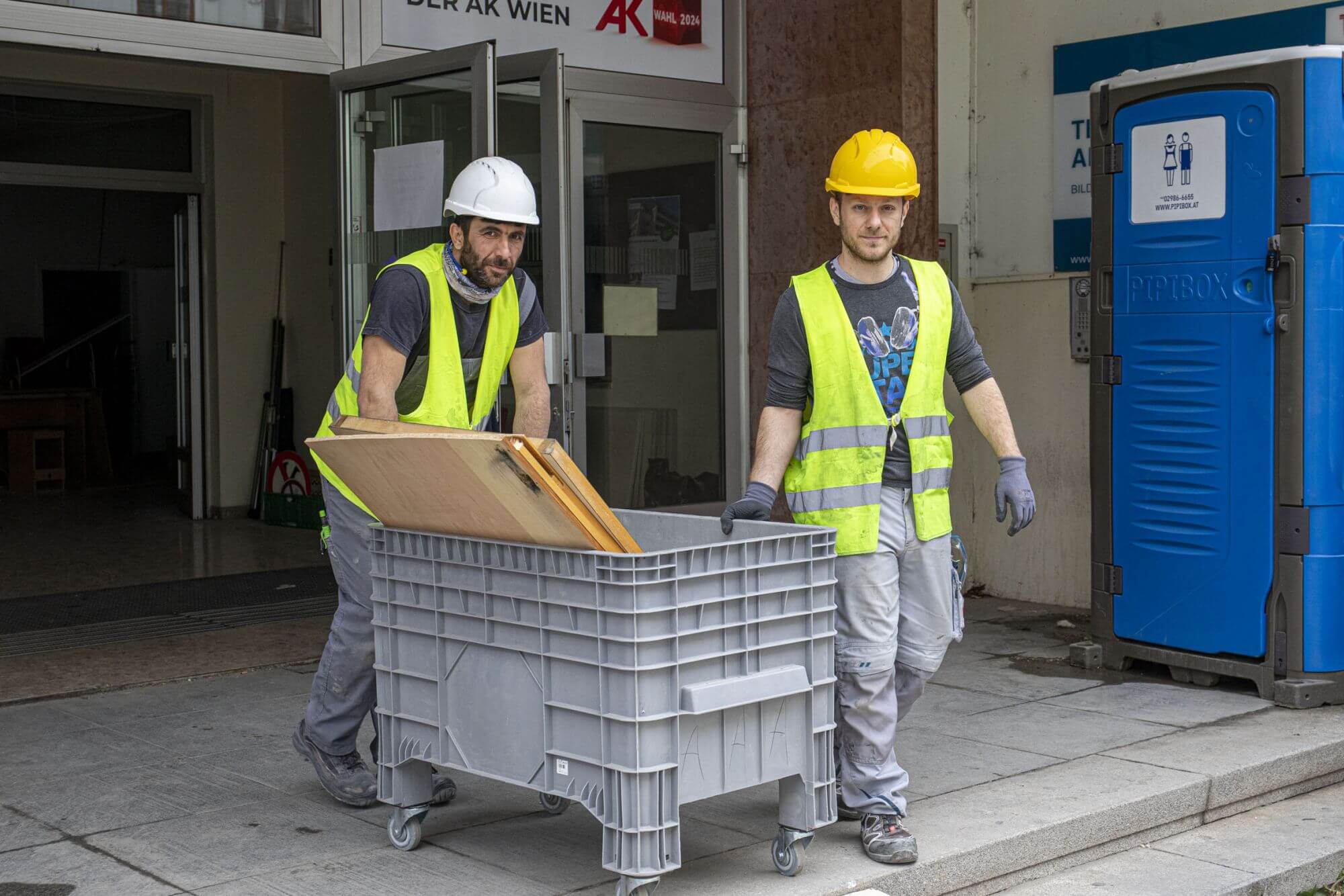 Zwei Personen auf einer Baustelle transportieren altes Baumaterial in eine Entsorgungstonne.